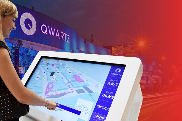 Woman using a ViaDirect interactive wayfinding kiosk displaying a shopping mall map at Qwartz shopping center in France.