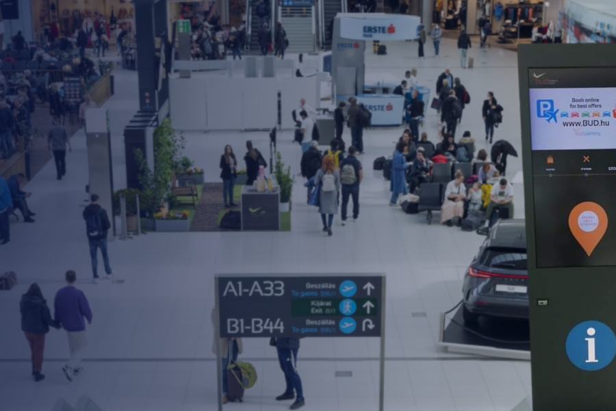 An interactive kiosk with 49-inch screens at Budapest Ferenc Liszt International Airport, providing real-time flight information and directions to boarding gates, located in the Airside area near the duty-free zone.