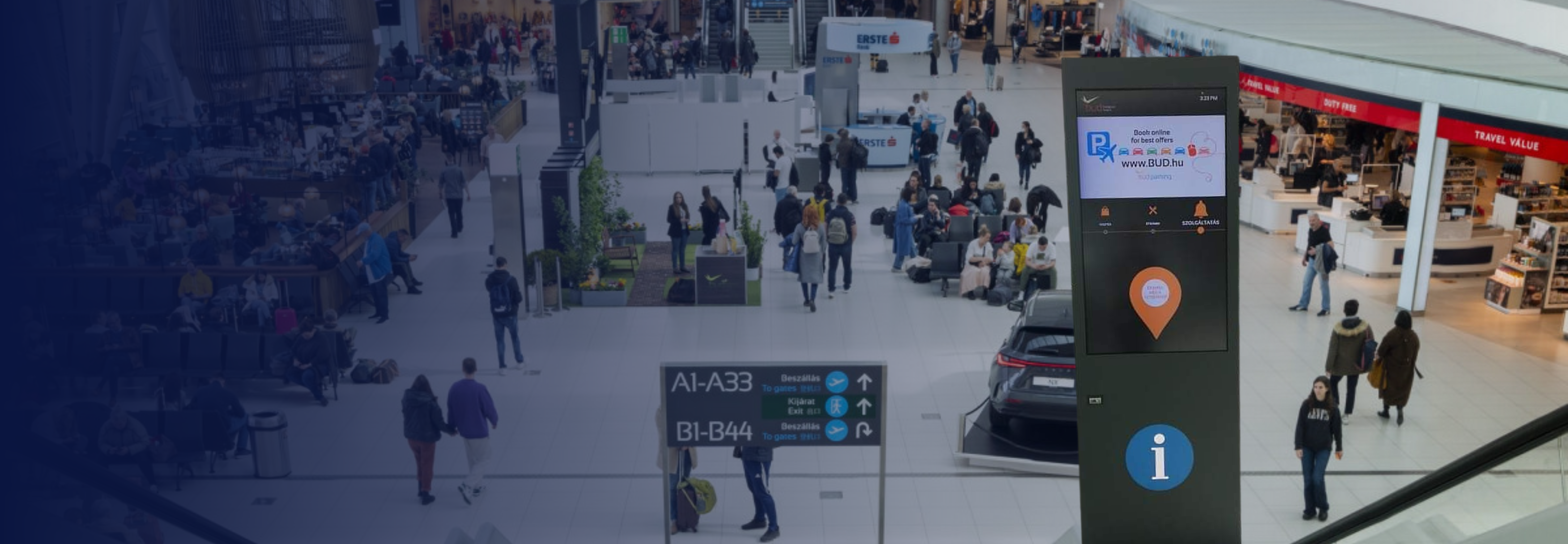 An interactive kiosk with 49-inch screens at Budapest Ferenc Liszt International Airport, providing real-time flight information and directions to boarding gates, located in the Airside area near the duty-free zone.